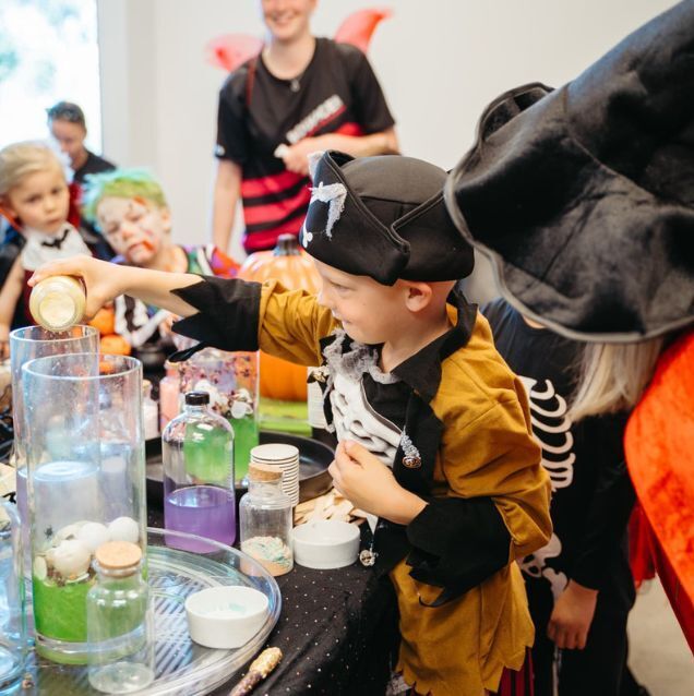 A young child dressed as a pirate excitedly pours a potion into a bubbling experiment at a themed party. Other costumed children watch in awe, participating in Sparkle & Spell Co.'s immersive magic potion party in Auckland.